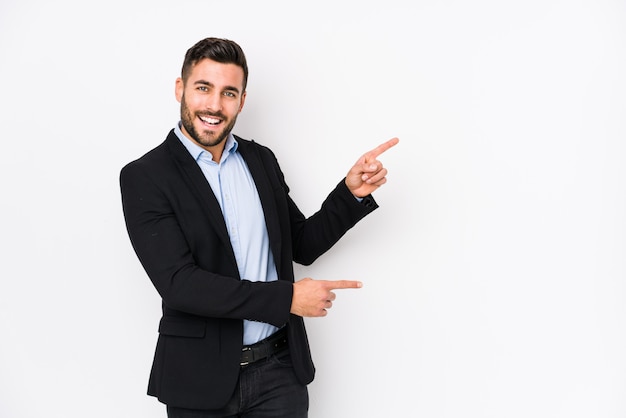 Jeune homme d'affaires caucasien contre un mur blanc isolé excité pointant avec les index loin.