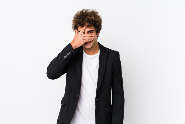 Jeune homme d'affaires caucasien sur un fond blanc isolé clignote à la caméra à travers les doigts, visage de couverture embarrassé.