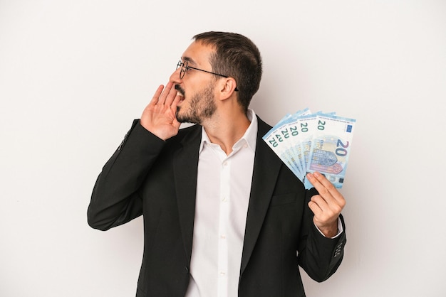 Jeune homme d'affaires caucasien tenant des billets de banque isolés sur fond blanc criant et tenant la paume près de la bouche ouverte