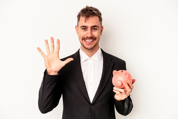 Jeune homme d'affaires caucasien tenant tirelire isolé sur fond blanc souriant joyeux montrant le numéro cinq avec les doigts.