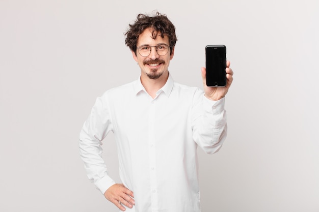 Jeune homme d'affaires avec une cellule souriant joyeusement avec une main sur la hanche et confiant