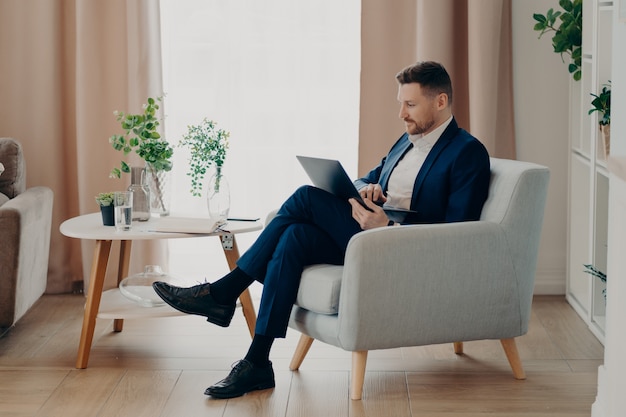 Jeune homme d'affaires concentré en costume élégant assis dans un fauteuil dans le salon d'un appartement moderne et regardant un écran d'ordinateur portable tout en travaillant à distance à la maison. Gens d'affaires et travail à distance