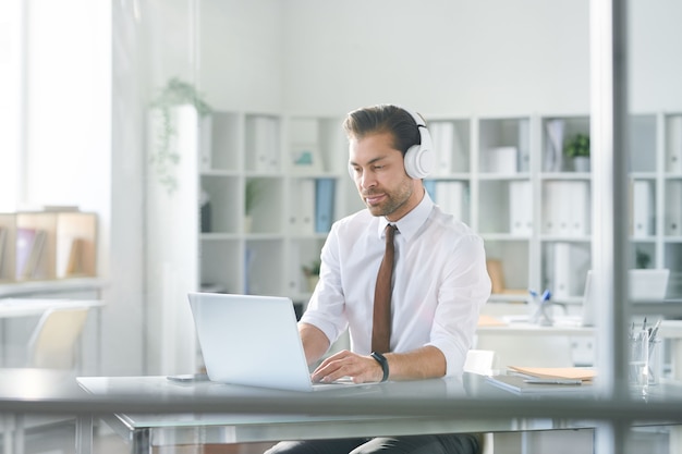 Jeune homme d'affaires concentré dans les écouteurs en tapant sur le clavier de l'ordinateur portable tout en regardant à travers les données en ligne