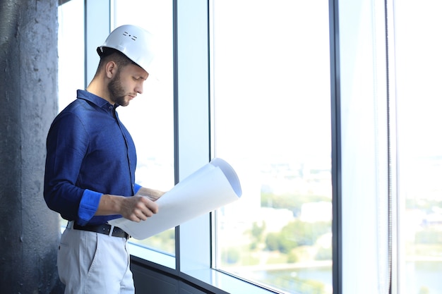 Jeune homme d'affaires confiant en chemise examinant le plan tout en se tenant contre une fenêtre au bureau.