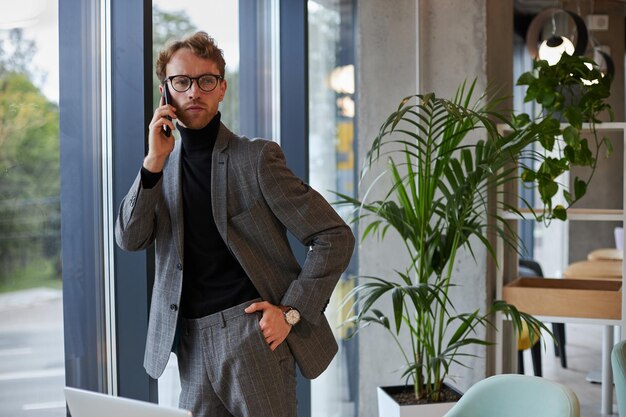 Jeune homme d'affaires confiant utilisant le téléphone pour négocier avec des investisseurs debout près de la fenêtre du bureau