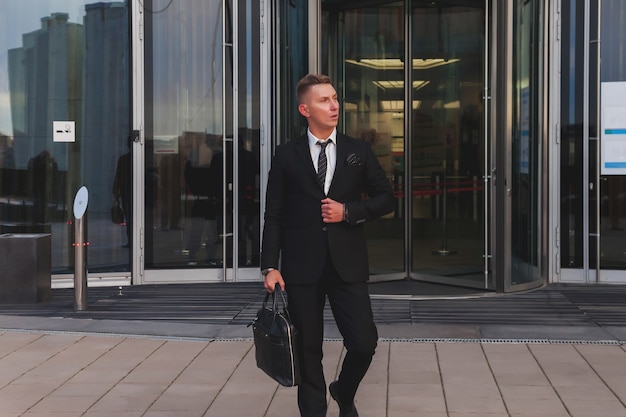 Un jeune homme d'affaires contemplatif portant des vêtements de style et avec une mallette quitte le bâtiment d'affaires près de l'entreprise, mode de vie