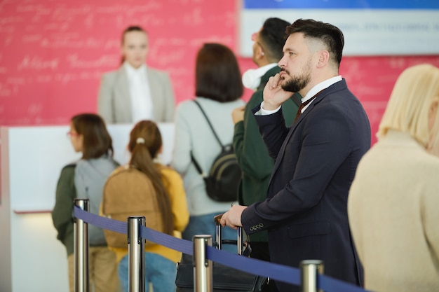 Jeune homme d'affaires en costume parlant au téléphone mobile tout en se tenant dans une file d'attente et en attendant son inscription