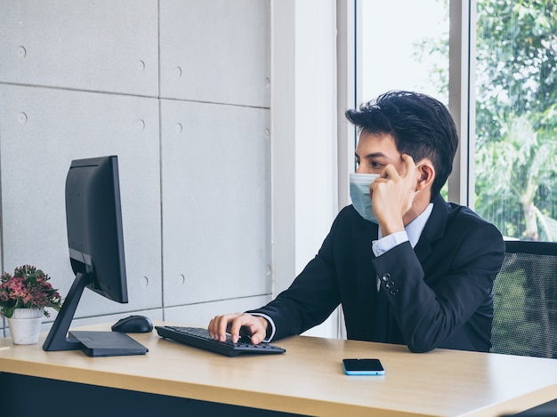 Jeune homme d'affaires en costume portant un masque protecteur travaillant sur ordinateur et pensant avec tension avec un visage fatigué avec un smartphone sur un bureau près d'une immense fenêtre en verre.