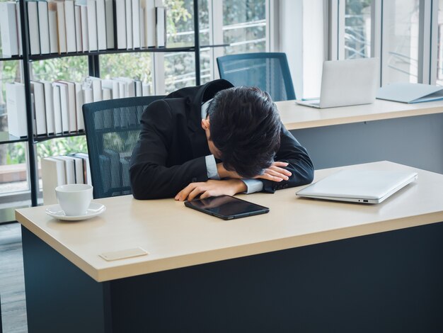 Jeune Homme D'affaires En Costume Avec Des Problèmes, Fatigué, Stressé Et Triste Ennuyeux Assis Avec La Tête Baissée Sur Son Bureau Au Bureau.