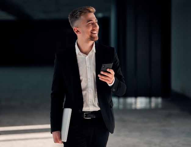 Un jeune homme d'affaires dans un centre d'affaires avec un téléphone et un ordinateur portable à la main dans un costume noir et une chemise blanche et souriant