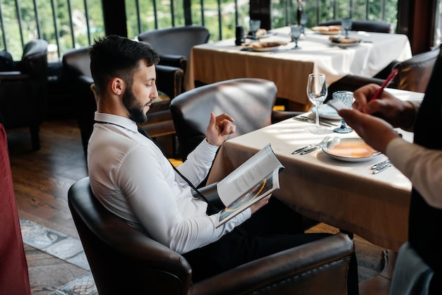 Un jeune homme d'affaires dans un restaurant raffiné examine le menu et passe une commande à un jeune serveur dans un tablier élégant Service client Service de table dans le restaurant