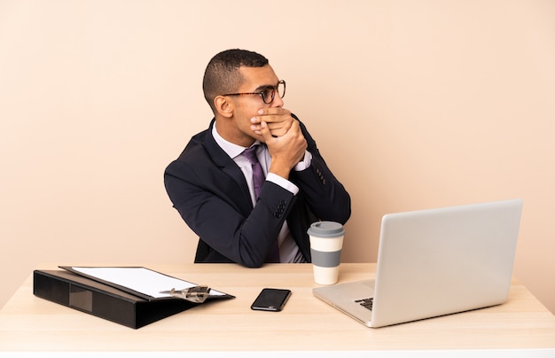 Jeune homme d'affaires dans son bureau avec un ordinateur portable et d'autres documents couvrant la bouche et regardant sur le côté