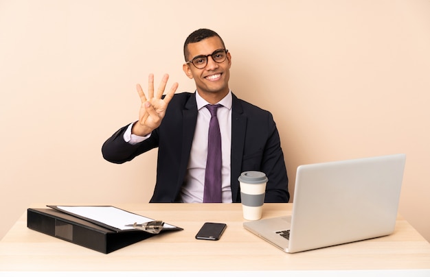 Jeune homme d'affaires dans son bureau avec un ordinateur portable et d'autres documents heureux et en comptant quatre avec les doigts