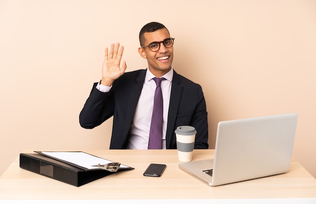 Jeune homme d'affaires dans son bureau avec un ordinateur portable et d'autres documents saluant avec la main avec une expression heureuse