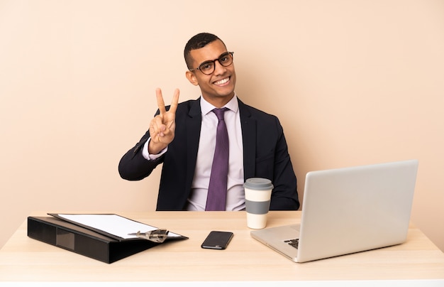 Jeune homme d'affaires dans son bureau avec un ordinateur portable et d'autres documents souriant et montrant le signe de la victoire