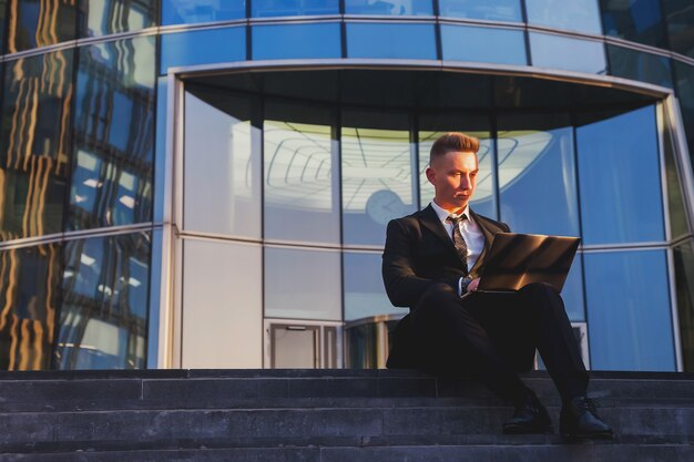 Jeune homme d'affaires dans le style porter des vêtements, costume assis sur les marches avec ordinateur portable soirée au coucher du soleil après le travail. Banquier masculin décontracté dans le bureau d'arrière-plan situé dans le quartier financier. Espace de copie