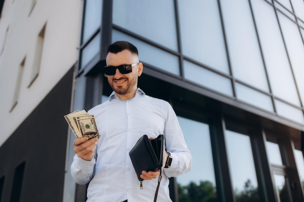 Jeune homme d'affaires debout appuyé sur le mur portant des lunettes dans la rue de la ville comptant de l'argent souriant joyeux