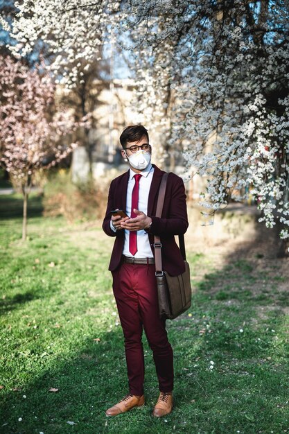 Jeune homme d'affaires debout dans un parc, utilisant un téléphone et portant un masque de protection. Soins de santé, protection contre les virus, concept de protection contre les allergies.