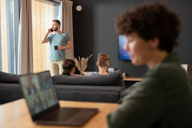 Jeune homme d'affaires décontracté en tenue décontractée debout près de la fenêtre, buvant et parlant au téléphone portable pendant que ses deux enfants regardaient la télévision