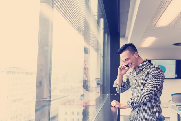jeune homme d'affaires détendu parlant sur un téléphone intelligent dans la salle de réunion du bureau d'affaires de démarrage moderne avec grande fenêtre et ville à backgronud