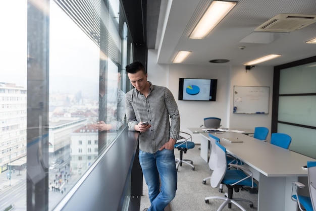 jeune homme d'affaires détendu utilisant un téléphone intelligent dans la salle de réunion du bureau d'affaires de démarrage moderne avec grande fenêtre et ville à backgronud