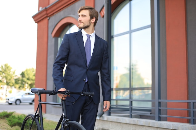 Jeune homme d'affaires élégant habillé en costume marchant avec un vélo dans une rue de la ville.