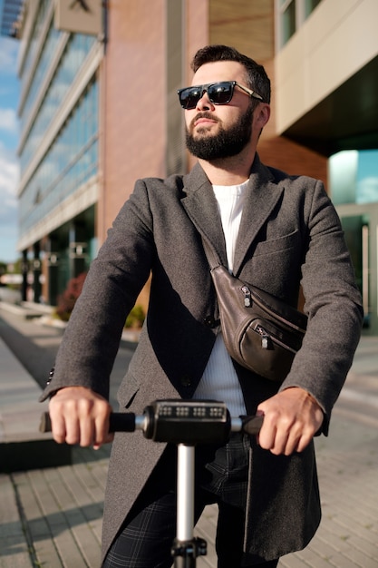 Jeune homme d'affaires élégant en manteau et lunettes de soleil à cheval sur un scooter électrique en milieu urbain contre un immeuble de bureaux moderne