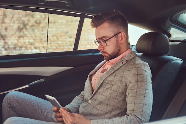 Jeune homme d'affaires élégant et prospère dans un costume gris et une chemise rose, à l'aide d'une tablette, à cheval sur un siège arrière dans une voiture de luxe.