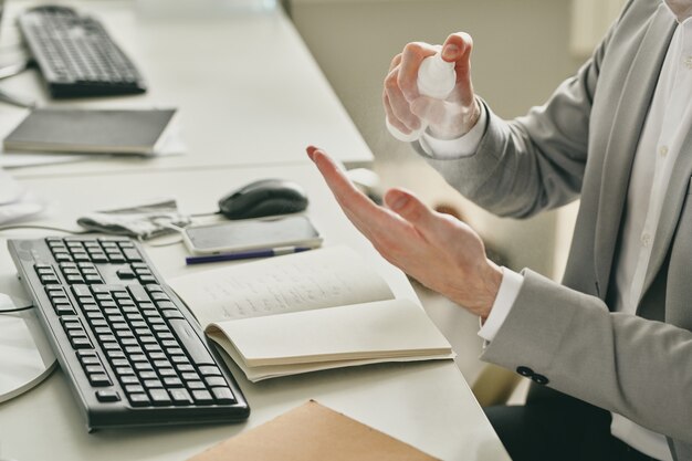 Jeune homme d'affaires élégant pulvérisant santitizer sur ses mains alors qu'il était assis par un bureau en face de l'ordinateur au bureau et va commencer à travailler