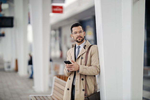 Un jeune homme d'affaires élégant s'appuie sur un pilier à la gare et attend le transport.