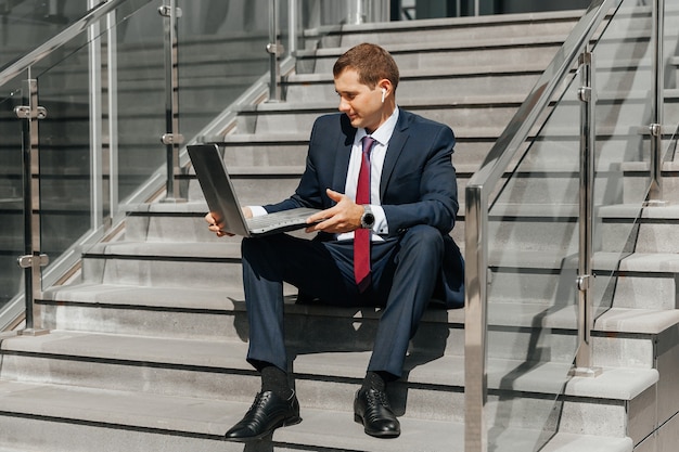 Un jeune homme d'affaires est assis dans les escaliers et travaille sur un ordinateur portable. Un jeune homme est assis sur les marches à l'extérieur et travaille sur un ordinateur portable.