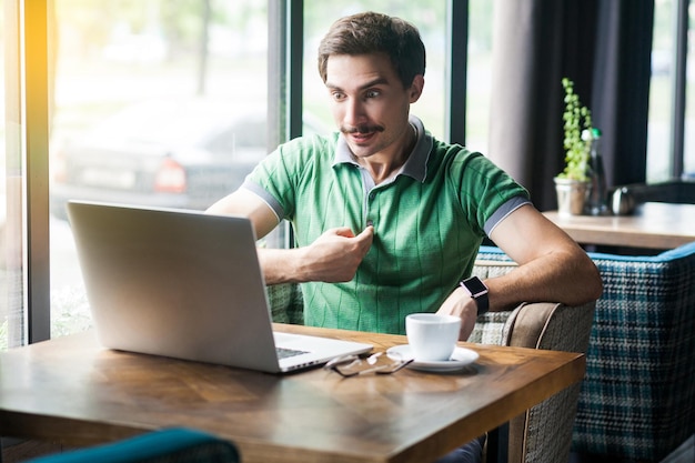 Jeune homme d'affaires étonné en t-shirt vert assis pointant et regardant l'écran de l'ordinateur portable avec un visage surpris demandant des affaires et un concept indépendant tourné en intérieur près d'une grande fenêtre pendant la journée