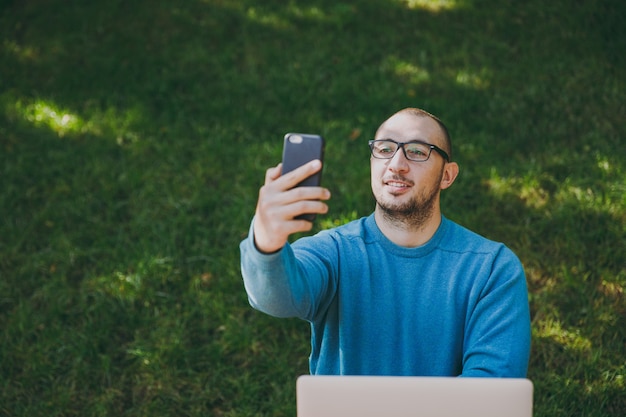 Jeune homme d'affaires ou étudiant intelligent et réussi en chemise bleue décontractée, lunettes assis à table faisant du selfie sur téléphone portable dans le parc de la ville à l'aide d'un ordinateur portable, travaillant à l'extérieur. Concept de bureau mobile.