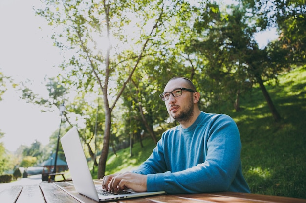 Jeune homme d'affaires ou étudiant intelligent réussi dans des lunettes de chemise bleue décontractée assis à table avec un téléphone portable dans le parc de la ville à l'aide d'un ordinateur portable travaillant à l'extérieur sur la nature verdoyante. Concept de bureau mobile.