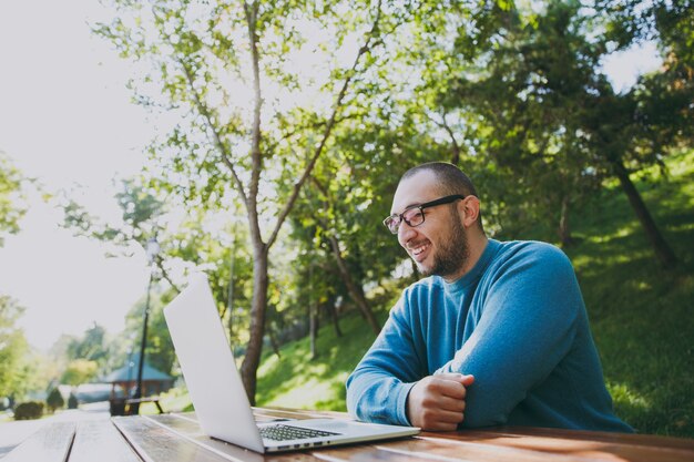 Jeune homme d'affaires ou étudiant intelligent réussi dans des lunettes de chemise bleue décontractée assis à table avec un téléphone portable dans le parc de la ville à l'aide d'un ordinateur portable travaillant à l'extérieur sur la nature verdoyante. Concept de bureau mobile.