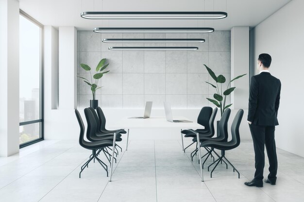 Jeune homme d'affaires européen debout dans une salle de réunion moderne en béton blanc avec fenêtre de meubles avec vue sur la ville et plantes décoratives