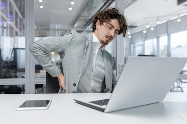 Photo jeune homme d'affaires fatigué assis à son bureau dans le bureau travaillant sur un ordinateur portable et tenant son