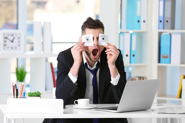 Jeune homme d'affaires avec de faux yeux peints sur des autocollants en papier bâillant sur le lieu de travail au bureau