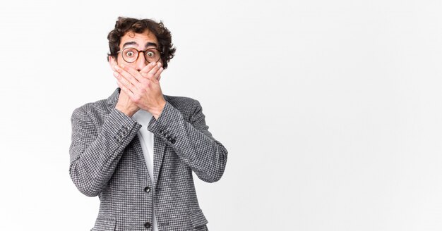 Jeune homme d'affaires fou couvrant la bouche avec les mains avec une expression choquée et surprise, gardant un secret ou disant oups contre le mur plat