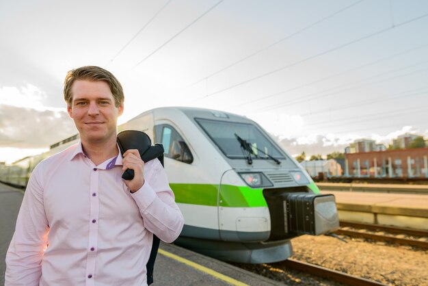 Jeune homme d'affaires heureux souriant contre le soleil devant le train à la gare