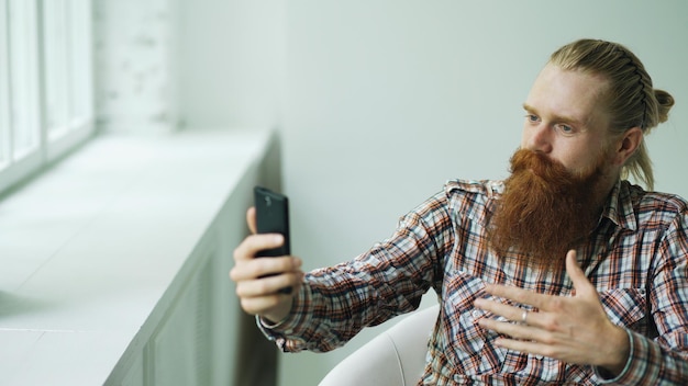 Jeune homme d'affaires hipster barbu parlant chat vidéo sur smartphone tout en étant assis dans une chaise de bureau