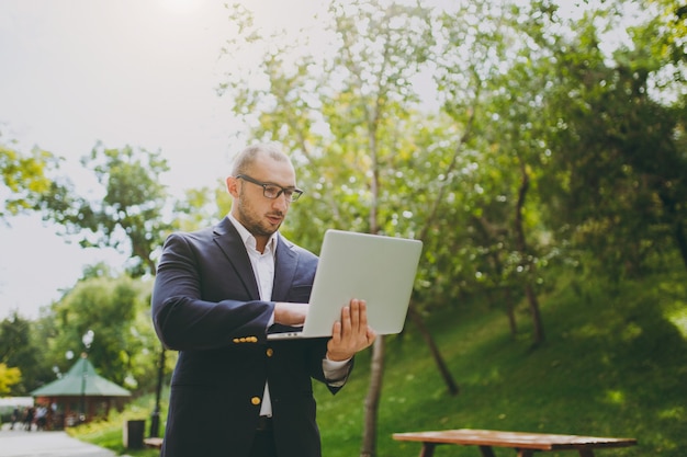 Jeune homme d'affaires intelligent réussi en chemise blanche, costume classique, lunettes. Homme debout et travaillant sur un ordinateur portable dans un parc de la ville à l'extérieur sur fond de nature. Bureau mobile, concept d'entreprise.
