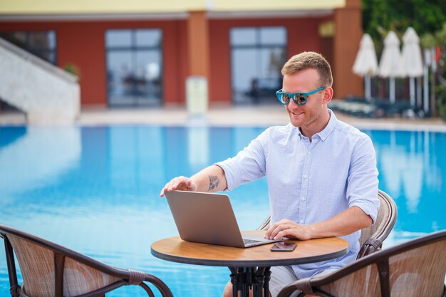 Un jeune homme d'affaires à lunettes de soleil travaille sur un ordinateur portable assis à une table près de la piscine. Travail à distance. Indépendant