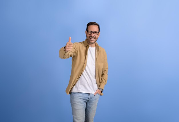 Jeune homme d'affaires avec la main dans la poche souriant et faisant des gestes avec le pouce en l'air contre un fond bleu