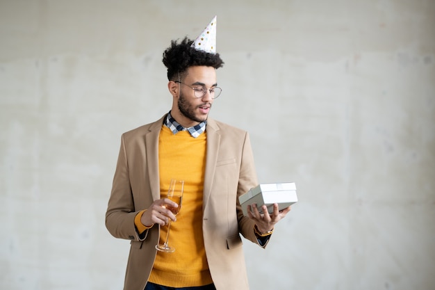 Jeune homme d'affaires métis en tenue décontractée et casquette d'anniversaire tenant une flûte de champagne pétillant tout en regardant une boîte-cadeau dans sa main