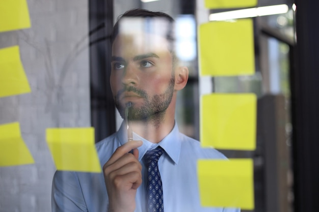 Jeune homme d'affaires moderne utilisant des notes adhésives tout en se tenant derrière le mur de verre du bureau.