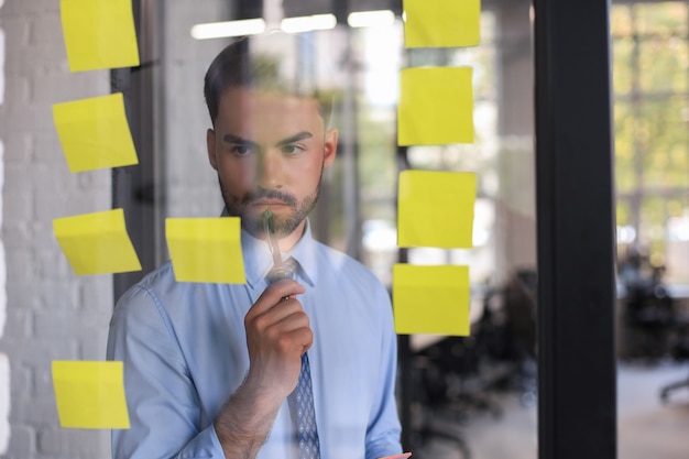 Jeune homme d'affaires moderne utilisant des notes adhésives tout en se tenant derrière le mur de verre du bureau.