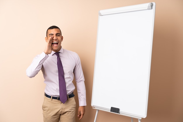 Jeune homme d'affaires sur un mur isolé donnant une présentation sur tableau blanc et criant avec la bouche grande ouverte