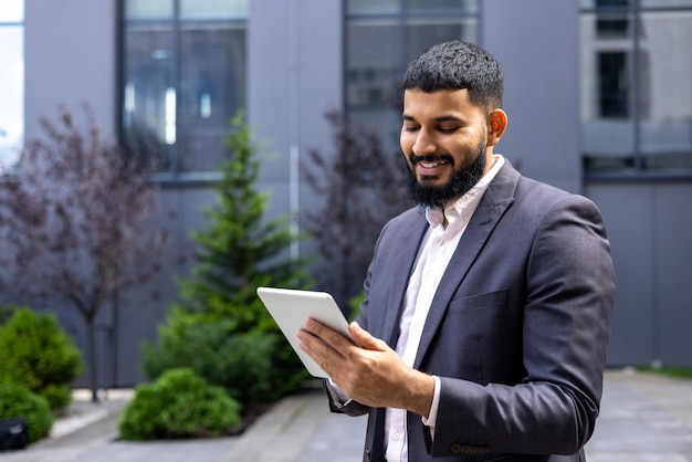 Jeune homme d'affaires musulman souriant, debout à l'extérieur du centre de bureau et utilisant une tablette en tapant un message