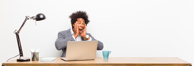 Jeune homme d'affaires noir se sentir heureux, excité et surpris, regardant sur le côté avec les deux mains sur le visage sur un bureau